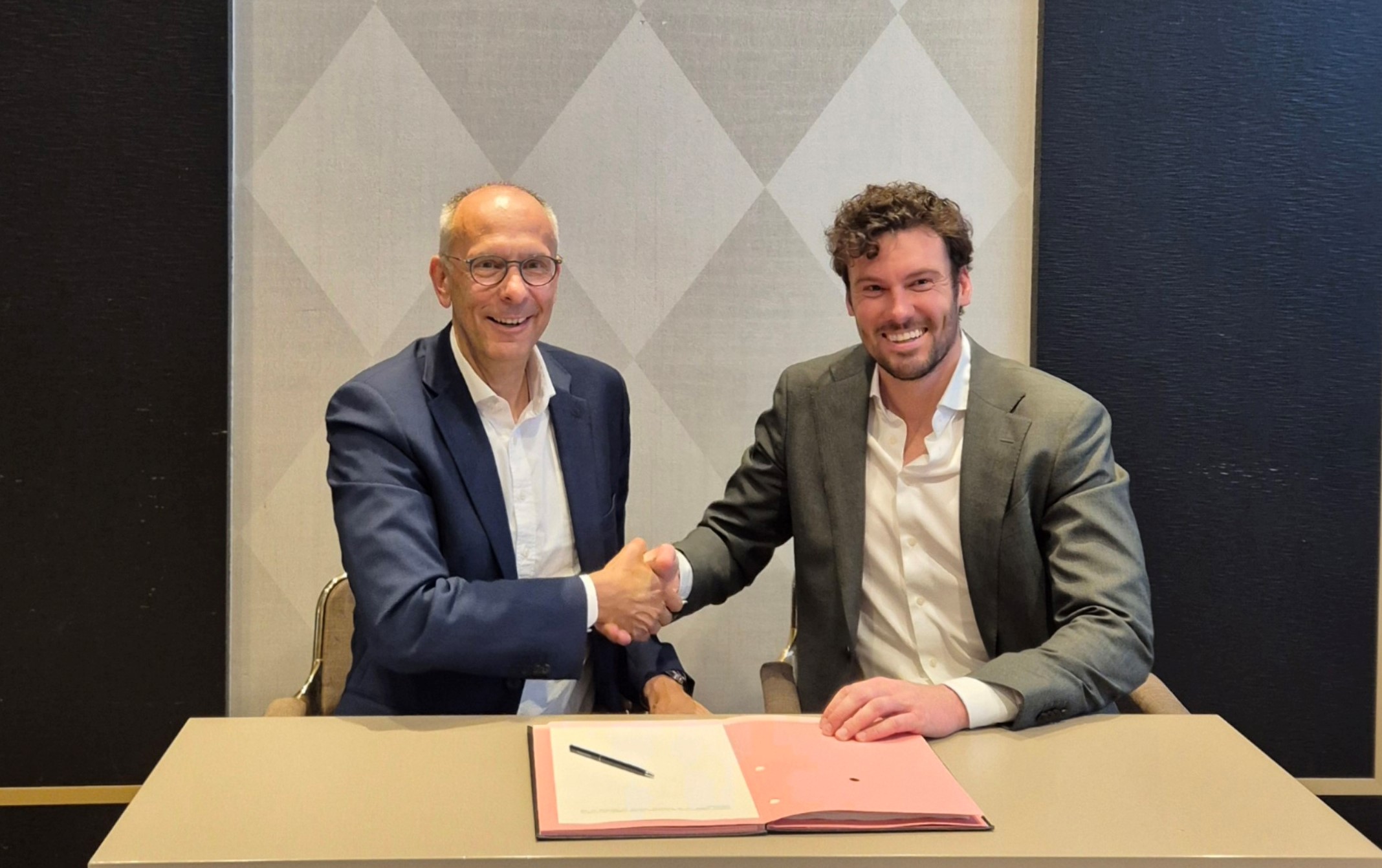 Stefan Falk und Jan Hedding beim Unterzeichnen der Vereinbarung. Beide lächeln in die Kamera während sie Hände schütteln.